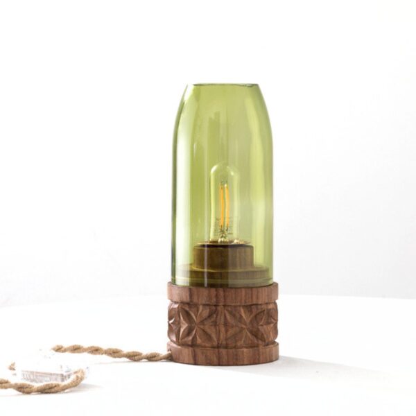 En bordlampe med grønn glasskjerm som trekker seg inn mot en åpen kant over pæren, stående på en trefot med mønster skåret rundt siden, med ledningen og bryteren ved siden av.