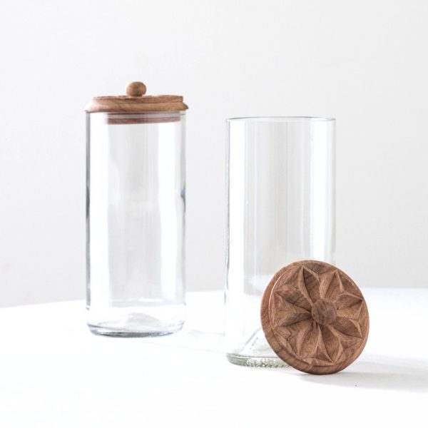 To høye klare glasskrukker med skårne trelokk, én lukket og én åpen med lokket lent mot seg.